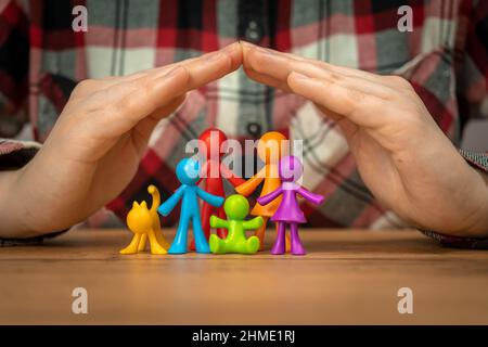 Familienversicherungskonzept mit bunten Familienfiguren, die mit Händen auf einem Holztisch bedeckt sind. Sicherheit und Unterstützung Stockfoto