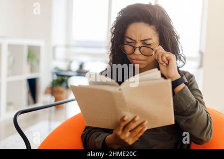 Fokussierte schwarze Dame in einer Brille versucht, Buch zu lesen Stockfoto