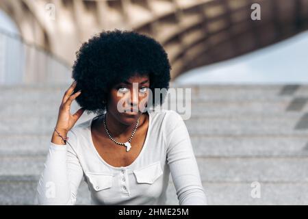 Porträt einer jungen schwarzen Frau, die auf einer Treppe auf der Straße sitzt und mit der Hand ihr afro Haar streichelte Stockfoto