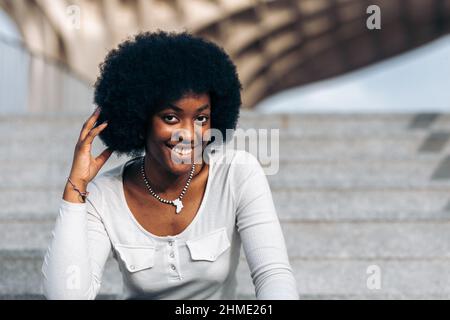 Porträt einer lächelnden jungen schwarzen Frau, die auf einer Treppe auf der Straße sitzt und mit der Hand über ihr afro Haar streichelt Stockfoto