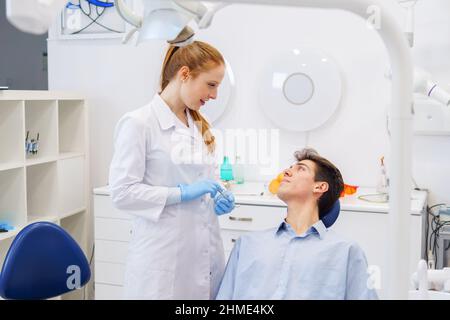 Junger Mann schaut auf Ingwer weibliche Arzt in medizinischen Uniform, während Sie zeitgenössische Zahnklinik besuchen Stockfoto