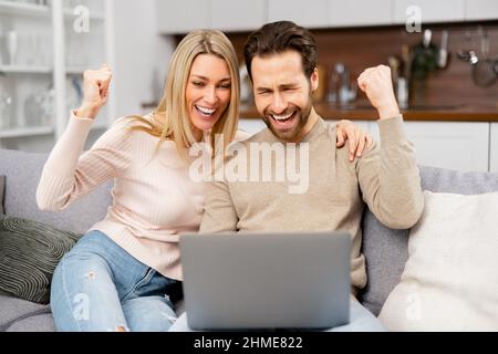 Ja. Fröhlicher Kerl bekam eine Beförderung bei der Arbeit, gewann die Lotterie, kaufte ein Urlaubsticket, sitzt am Laptop. Die kaukasische Frau und ihr Mann vor dem Laptop und freuen sich über ihren Erfolg Stockfoto