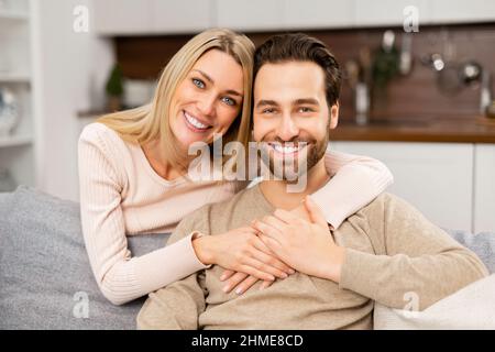 Porträt von schönen Paar zu Hause. Hübscher Mann, der auf der bequemen Couch sitzt, schöne blonde Frau, die sich von hinten umarmt. Ehepartner verbringen Zeit miteinander Stockfoto