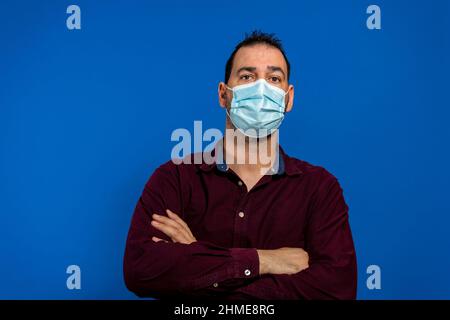 Porträt eines hübschen jungen Mannes in einer chirurgischen medizinischen Maske, gekleidet in einem violetten Hemd, in dem seine Arme isoliert auf blauem Hintergrund gekreuzt sind, innen Stockfoto