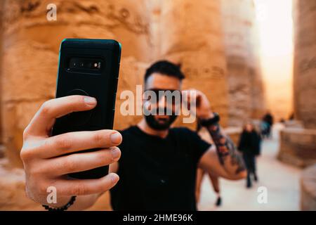 Hispanic Mann trägt eine Sonnenbrille und nimmt ein Selfie in luxor, umgeben von alten ägyptischen Säulen. Konzentrieren Sie sich auf das Smartphone, Reisende, die Fotos machen Stockfoto