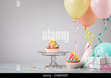 Zwei Rainbow Birthday Cupcakes mit Geschenken, Hüten und bunten Ballons auf hellgrauem Hintergrund. Szene von einer Geburtstagsfeier. Stockfoto