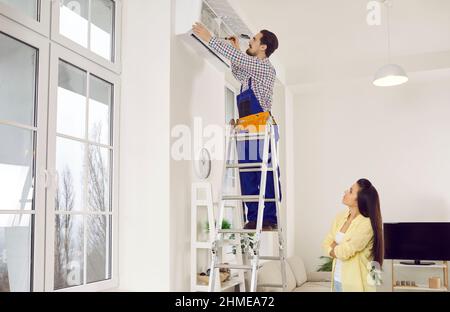 Techniker, der im Haus einer jungen Frau auf einer Leiter steht und die Klimaanlage an der Wand repariert Stockfoto