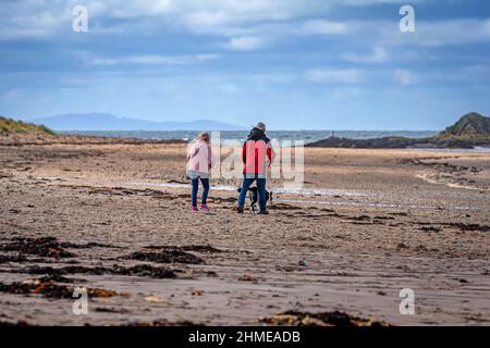 Paar, das einen Hund am Rhoseigr Strand an der Anglesey Küste, Anglesey, Wales, Großbritannien, spazieren geht Stockfoto