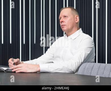Ein reifer Geschäftsmann im Trikot hört SMB zu, macht Notizen im Notizbuch, ein Planer am Konferenzschalter. Hochwertige Fotos Stockfoto