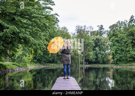 Frau mit gelbem Regenschirm, die am Pier am Teich steht. Sich änderndes Frühlingswetter in der Natur Stockfoto