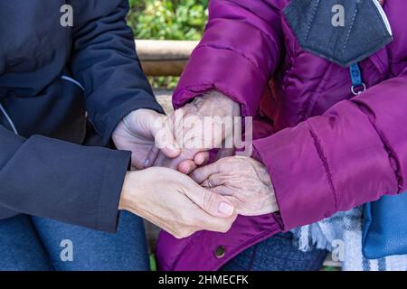 Eine reife Frau hält die Hände ihrer älteren Mutter, während sie auf einer Bank sitzt Stockfoto