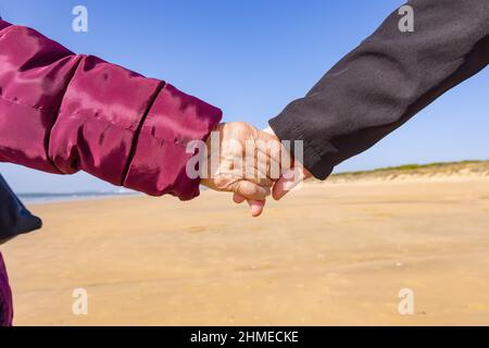 Eine reife Frau hält die Hand ihrer älteren Mutter, während sie am Strand spazieren geht Stockfoto