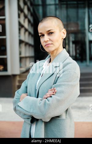 Selbstbewusste Geschäftsfrau, die die Kamera anschaut, während sie mit gekreuzten Armen im Freien posiert. Stockfoto