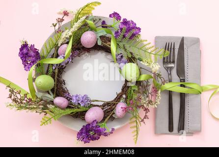 Ostertisch. Pastellfarbener Kranz mit Ei- und Blumendekoration auf rosa Hintergrund, Draufsicht. Frühlingsfeiertage. Stockfoto
