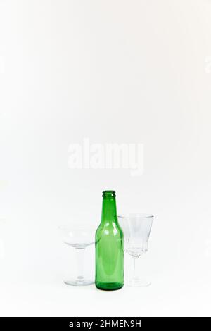 Set aus leerer Bierflasche aus grünem Glas mit zwei ebenso leeren transparenten Glaskelchen auf weißem Hintergrund. Copyspace. Vertikale Fotografie. Stockfoto