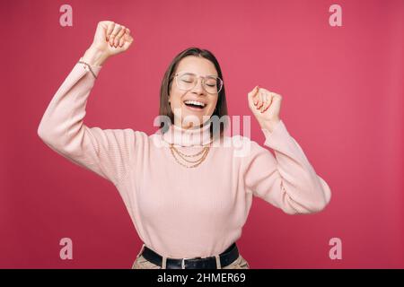 Aufgeregte junge Frau, die eine Brille mit geschlossenen Augen trägt, tanzt in einem Studio über einem rosa Hintergrund. Stockfoto
