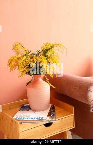 Vase mit Mimosenblumen und Zeitschriften auf dem Tisch im Innenraum des Zimmers Stockfoto