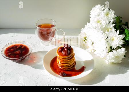 Fritters in einem weißen Teller mit Erdbeermarmelade, eine Tasse Tee und ein Bouquet von weißen Chrysanthemen auf einem weißen Tisch. Stockfoto