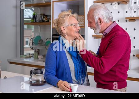 Ein glückliches älteres Paar in der Küche. Der Mann küsst seine Frau und dankt ihr für den Morgenkaffee. Stockfoto
