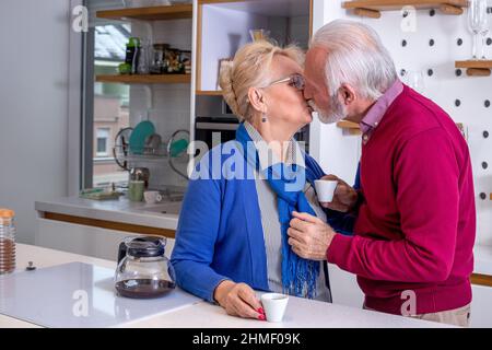 Ein glückliches älteres Paar in der Küche. Der Mann küsst seine Frau und dankt ihr für den Morgenkaffee. Stockfoto