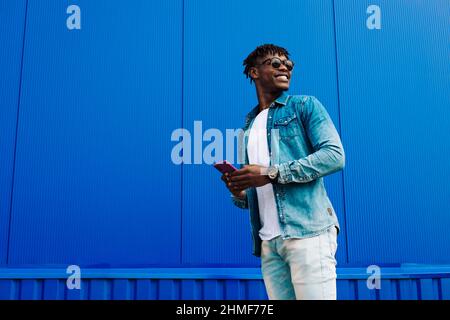 Die ganze Länge eines fröhlichen stilvollen afroamerikanischen Mannes in vollem Wachstum, nutzt ein Smartphone und geht an einer blauen Wand vorbei, während er zur Arbeit geht. Der Mann hält Stockfoto
