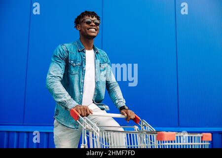 Stilvoller amerikanischer Mann mit Sonnenbrille, beim Laufen mit Einkaufswagen und lächelnd vor der Kamera. Ein gutaussehender Mann kauft in der Stadt ein und lächelt, während wa Stockfoto