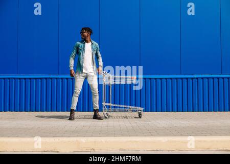 Schöner afroamerikanischer Mann, der im Push-Cart-Supermarkt einkauft. Seitenansicht eines fröhlichen jungen Mannes, mit einem Einkaufswagen, über einer blauen Wand mit Polizist Stockfoto