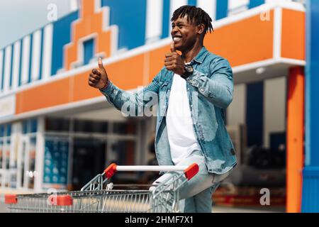 Mann beim Einkaufen. Lächelnder, glücklicher afrikanischer Mann mit Sonnenbrille, der aus dem Einkaufszentrum mit dem Daumen nach oben geht, Mann, der gerne einkaufen geht. Konsu Stockfoto