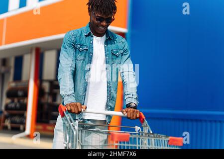 Mann beim Einkaufen. Lächelnder, fröhlicher dunkelhäutiger Mann mit Sonnenbrille verlässt das Einkaufszentrum mit einem Einkaufswagen, der die Straße entlang läuft. Konsum, Lifestyle konz Stockfoto