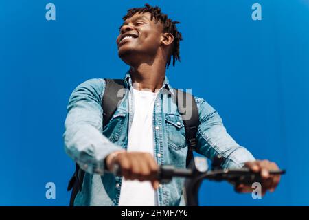 Glücklich lächelnd lockiger afroamerikanischer Mann in lässiger Kleidung, der an einem sonnigen Tag einen Elektroroller auf der Straße reitet.Freudvoller hübscher Mann, der einen Sco reitet Stockfoto