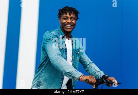 Glücklich lächelnd lockiger afroamerikanischer Mann in lässiger Kleidung, der an einem sonnigen Tag einen Elektroroller auf der Straße reitet.Freudvoller hübscher Mann, der einen Sco reitet Stockfoto