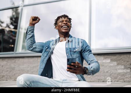Aufgeregt überglücklich afroamerikanischen Mann, mit Smartphone, Mann mit erhobenen Händen, feiert den Sieg, stehen vor dem Bürogebäude Stockfoto