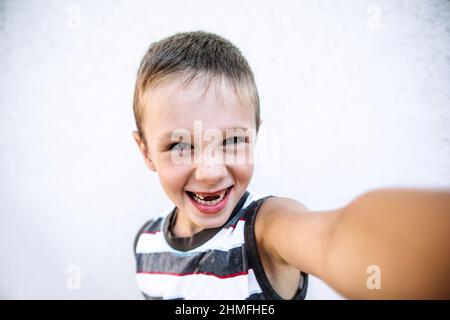 Junge mit Sommersprossen ohne zwei Zähne in legerer Kleidung macht Selfie an der Hand, Junge schaut in die Kamera und lächelt Stockfoto