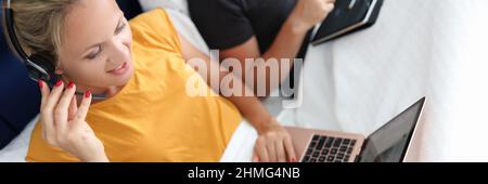 Eine junge Frau mit Kopfhörern und Mikrofon arbeitet im Bett auf dem Laptop Stockfoto