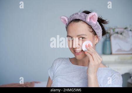 Close-up Kopfschuss lächelnd schöne Frau in ein Handtuch gewickelt, die Toner mit einem Wattestäbchen auf ihre Gesichtshaut nach einem Bad aufbrachte. Glückliche junge Dame, die morgens nach der Dusche die Hautpflege macht. Stockfoto