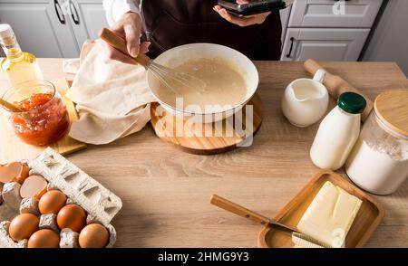 Die Hand einer Frau mit einem Schneebesen schlägt den Teig in einer Schüssel. Die andere Hand hält das Telefon mit dem Rezept für das weitere Kochen Stockfoto