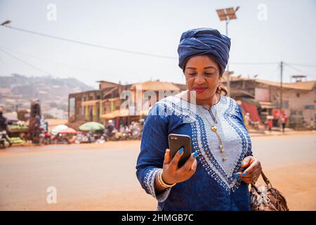 Eine afrikanische Frau in einem traditionellen Kleid sieht auf den Bildschirm ihres Smartphones. Die 50-jährige Frau in einem afrikanischen Dorf nutzt Technologie Stockfoto