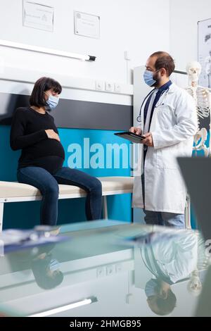 Geburtshelferin mit Tablette, die der Schwangeren medizinische Ratschläge gibt und Gesundheitsberatung im Büro macht. Werdende Patientin, die vom Arzt mit Gesichtsmaske Unterstützung bei der Mutterschaft erhält. Stockfoto