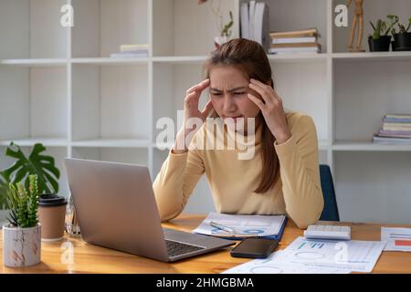 Die arbeitende Frau hat Kopfschmerzen, die junge asiatische Geschäftsfrau, die an einem Laptop arbeitet und Papierkram betont wird, hat Kopfschmerzen und denkt hart im Büro Stockfoto