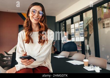 Glückliche junge Geschäftsfrau lächelt an der Kamera, während sie ein Smartphone hält. Fröhliche junge Geschäftsfrau, die mit ihrem Kollegen auf einem Sitzungstisch sitzt Stockfoto