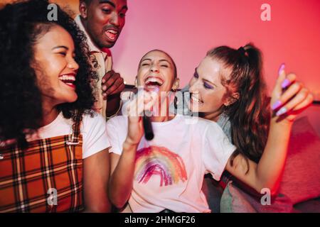 Fröhliche Freunde singen ihr Lieblingslied während der Karaoke-Nacht. Fröhliche junge Freunde, die bei einer Hausfeier fröhlich lachen. Gruppe von multikulturellen Freunden Stockfoto
