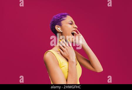 Zu meinen Lieblingsliedern mitsingen. Unbeschwerte junge Frau, die Musik über ihre kabellosen Kopfhörer hört. Frau mit purpurnen Haaren, die laut agains singt Stockfoto