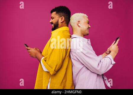 Lächelndes, schwules Paar, das in einem Studio Mobiltelefone benutzt. Glückliches junges Paar, das vor einem violetten Hintergrund zurück an den Rücken steht und SMS schreibt. Süßes Schwulencoupl Stockfoto