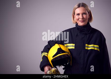Studio Portrait Of Smiling Reife Feuerwehrfrau Mit Helm Vor Schlichem Hintergrund Stockfoto