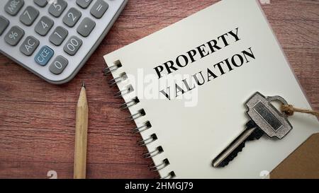 Text zur Immobilienbewertung auf Notizbuch mit Taschenrechner, Schlüssel, Bleistift und braunem Holzschreibtischhintergrund. Stockfoto