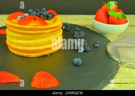 Stapel hausgemachter Pfannkuchen zum Frühstück. Heidelbeeren und Erdbeeren auf Pfannkuchen. Schwarzer Hintergrund, Draufsicht. Stockfoto