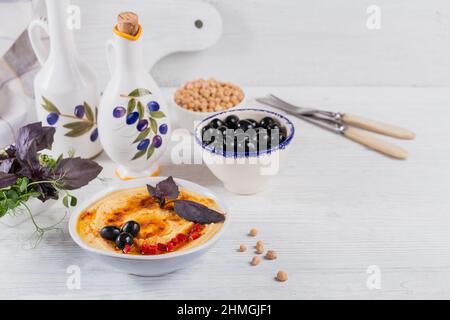 Eine Schüssel mit Kichererbsen hausgemachten Hummus mit Olivenöl, Basilikum, schwarzen Oliven und geräuchertem Paprika auf weißem Holzhintergrund. Stockfoto