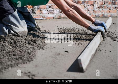 Nahaufnahme eines Baumeisters, der auf der Baustelle auf dem Dach eine Estrich-Schiene auf den mit Sand-Zement-Gemisch überzogenen Boden legte. Männlicher Arbeiter nivellieren Oberfläche mit gerader Kante beim Estrichboden. Stockfoto