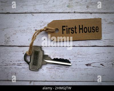 Draufsicht des Textes - erstes Haus auf braunem Schlüsselbund. Weißer Schreibtisch im Hintergrund. Stockfoto