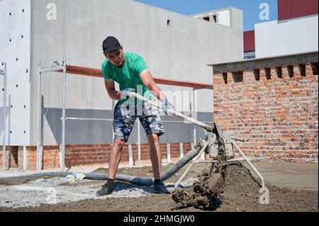 Männlicher Arbeiter schaufelt Sand-Zement-Mischung während der Arbeit auf der Baustelle. Mann bereitet Bodenestrich vor, während er im Freien in der Nähe der Betonestrich-Mischmaschine arbeitete. Stockfoto